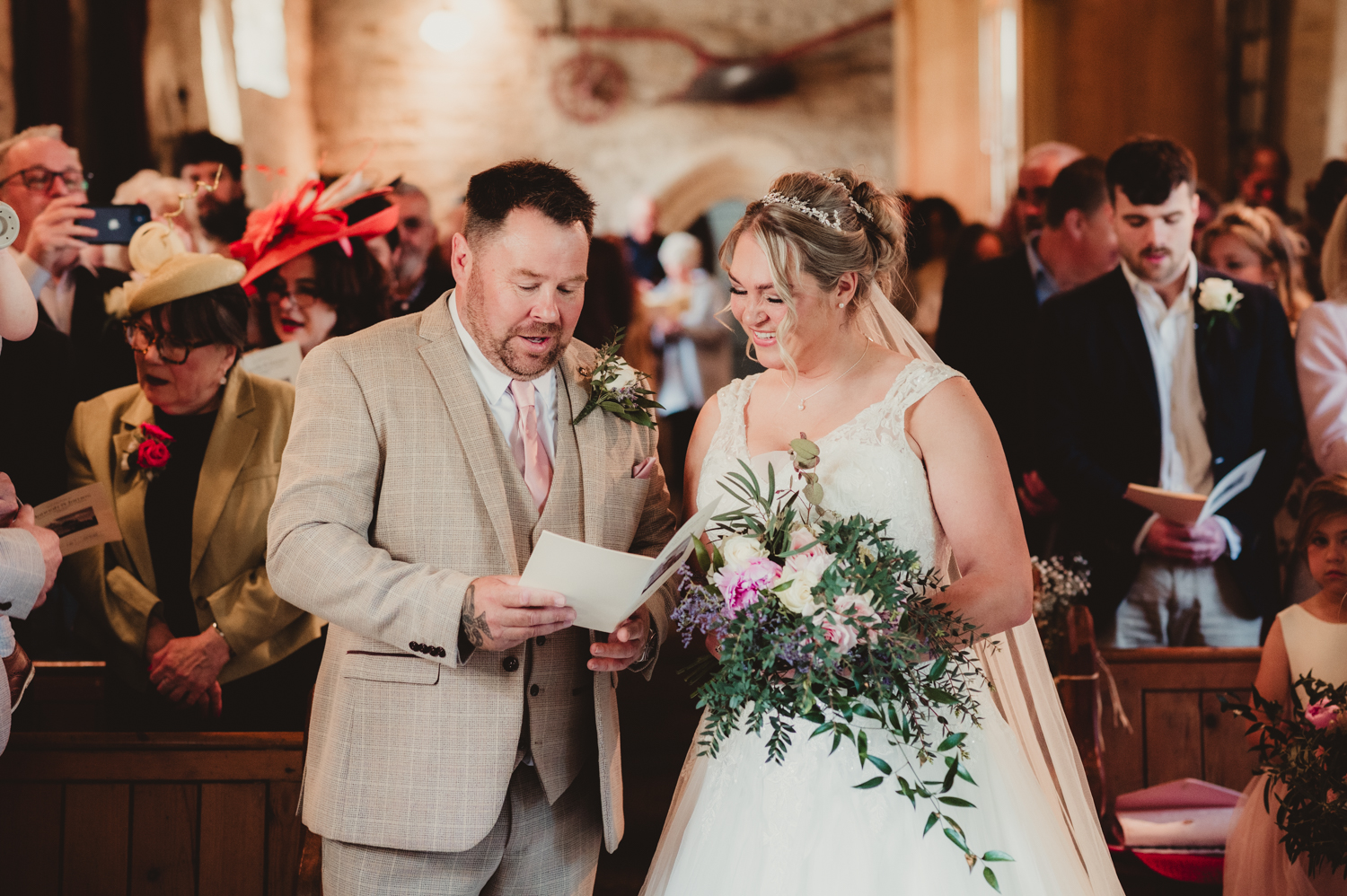 Bride and groom smiling and holding hymn sheets during their church ceremony, surrounded by family and friends at Boddington Church