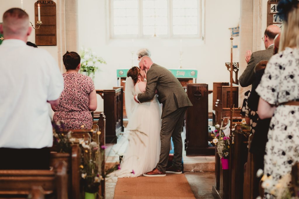 A bride and groom sharing their first kiss at St Marys Church, Bishopstone. A beautiful cotswolds wedding.