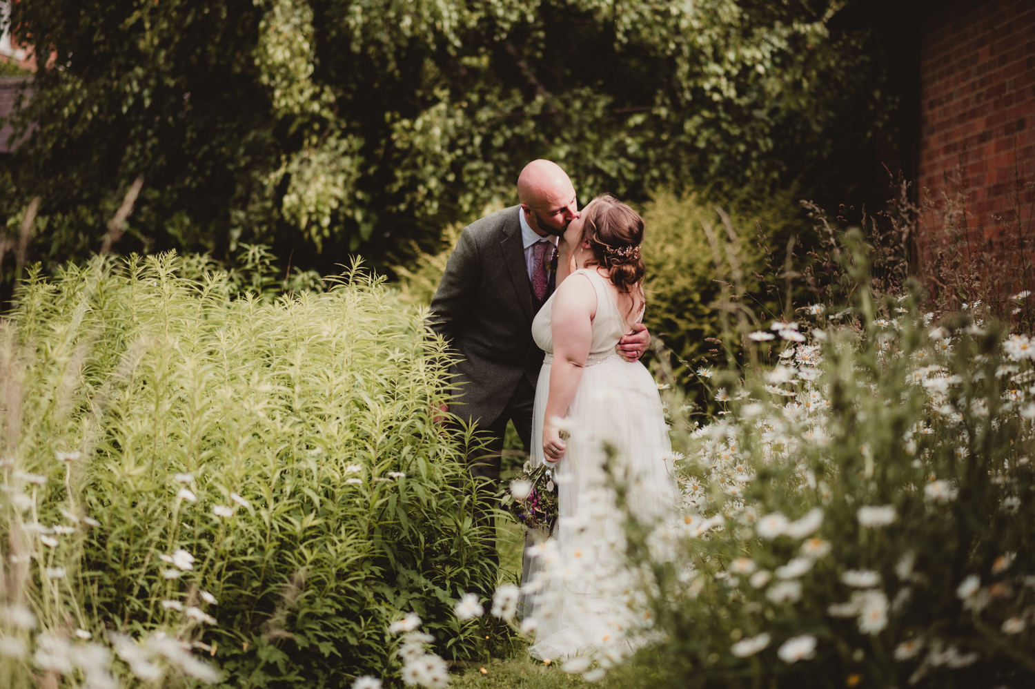 A bride and groom share a tender kiss in a lush garden setting. Surrounded by tall greenery and wildflowers, the moment feels intimate and natural. The bride, dressed in a flowing white gown, holds a small bouquet, while the groom, wearing a grey suit, gently embraces her. The backdrop of trees and blooming daisies adds to the relaxed, romantic atmosphere of this Cotswolds wedding scene.