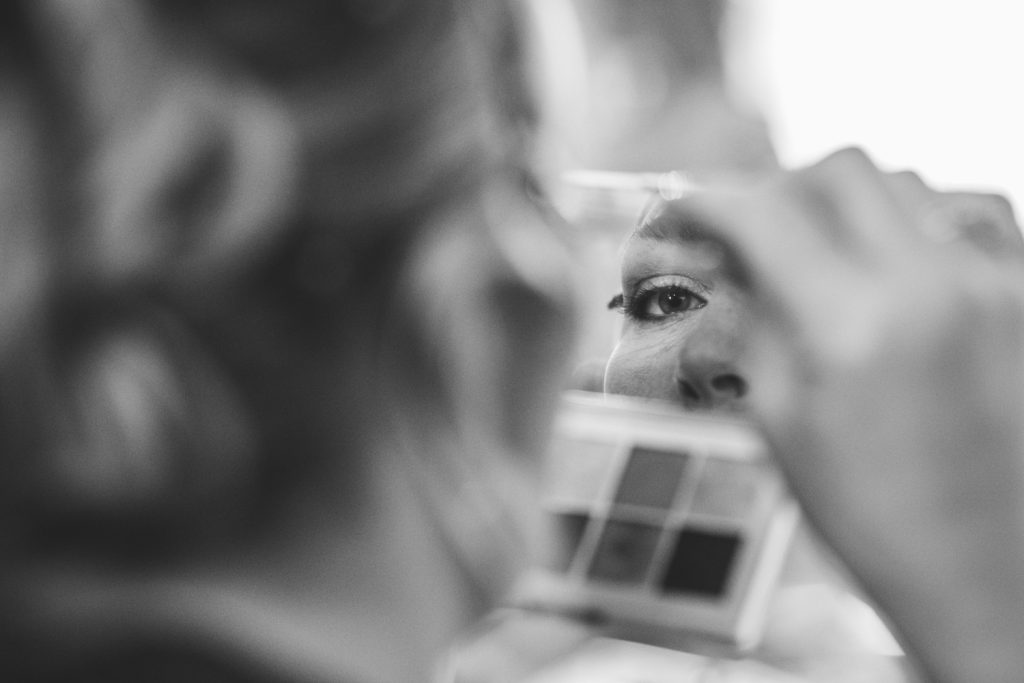 Close-up black and white shot of the bride applying makeup, with her reflection visible in a small mirror