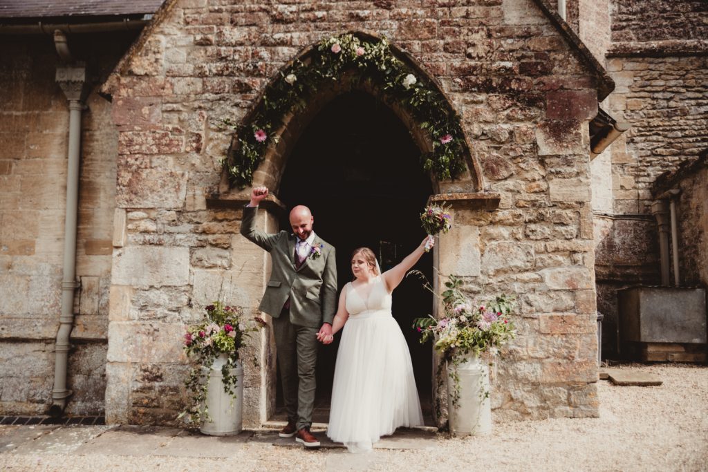 A bride and groom cheering as they exit St Marys Church in Bishopstone in the Cotswolds