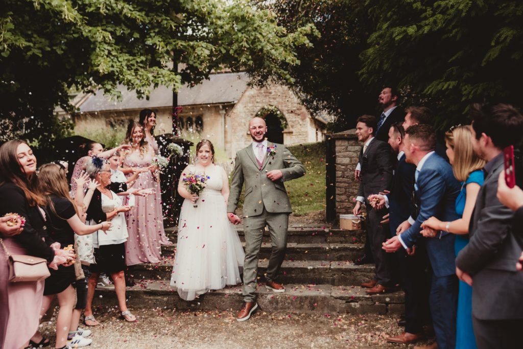 Bride & Groom step out of St Marys church in Bishopstone hand in hand, beaming as they are showered with confetti by their friends and family. A laid-back atmosphere of this beautiful Cotswolds countryside wedding.