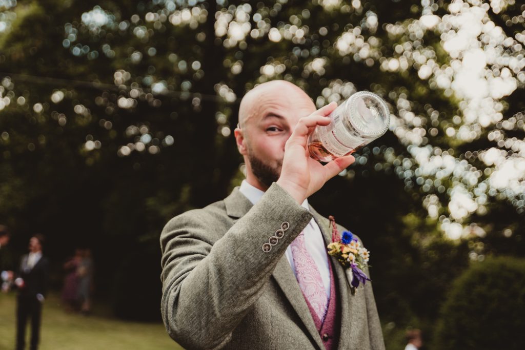 A candid photo of a groom downing his bottle of wine at his Cotswolds wedding