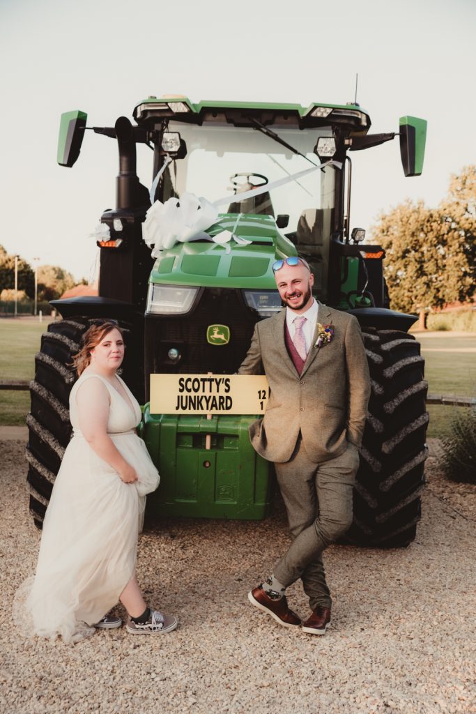 A newlywed couple poses next to a green John Deere tractor decorated with white ribbon, adding a fun farm twist to their wedding day. The groom, dressed in a grey suit and purple tie, leans casually against the tractor with a cheeky grin, while the bride, wearing a flowing white gown and Converse trainers, stands beside him with a relaxed, playful expression. A custom sign reading "Scotty's Junkyard" adds a quirky, personalised touch, capturing the couple’s love for farming and vintage vibes at their Cotswolds wedding.
