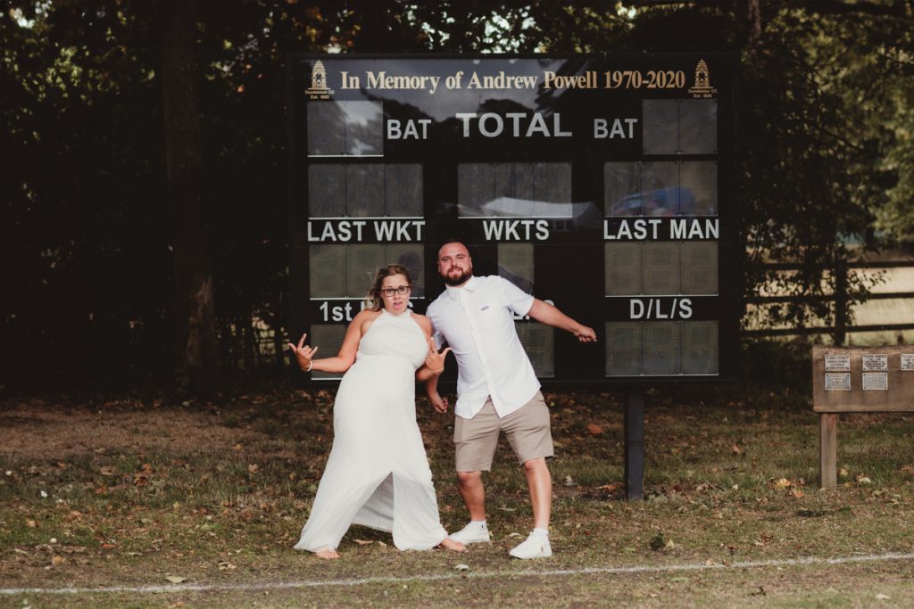 bride and groom in front of the cricket score board at Dumbleton Cricket Club